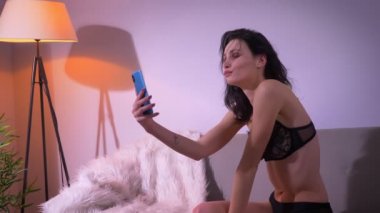 Siyah iç çamaşırı şehvetli esmer model kanepede oturan ve ev atmosferinde akıllı telefon selfies yapma.