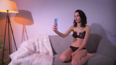 Siyah iç çamaşırı oyuncu esmer model kanepede oturan ve evde akıllı telefon komik yüzleri ile selfies yapma.