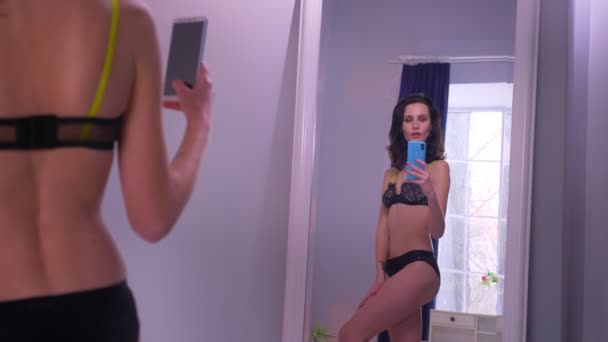 Mirror reflection of pretty and sexy brunette model in black lingerie posing to make photos at home. — Stock Video