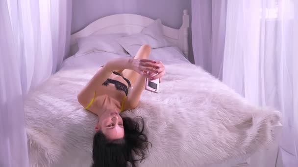 Sexy morena modelo em lingerie preta relaxante na cama dossel faz belas selfie-fotos no smartphone em casa . — Vídeo de Stock