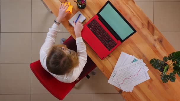 Close-up top shoot de jovem mulher de negócios caucasiana usando o laptop e escrever um lembrete sobre uma reunião nomeada dentro do escritório — Vídeo de Stock