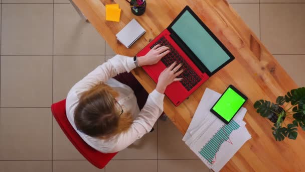 Close-up top shoot de jovem mulher de negócios caucasiana usando o laptop com o tablet com tela verde deitado na mesa dentro de casa no escritório — Vídeo de Stock