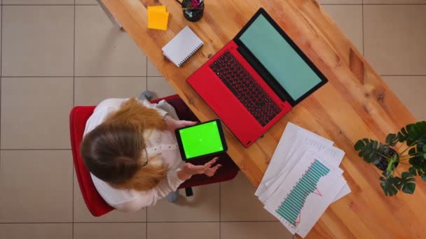 Primer plano brote superior de la joven mujer de negocios caucásica utilizando la tableta con pantalla verde sentado delante de la computadora portátil en el interior de la oficina — Vídeos de Stock