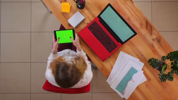 Close-up tiro superior de jovem mulher de negócios caucasiana mensagens de texto no tablet com tela verde sentado na frente do laptop dentro de casa no escritório — Vídeo de Stock