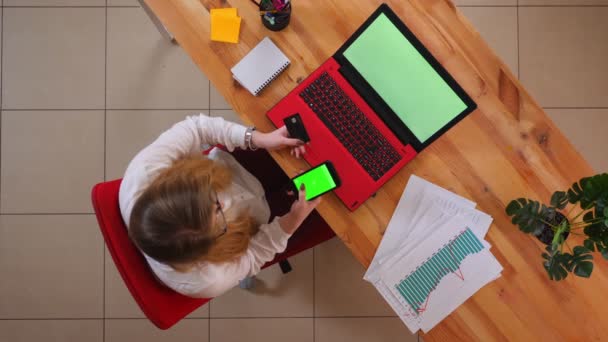 Primo piano top shot di giovane donna d'affari caucasica shopping online al telefono con schermo verde seduto di fronte al computer portatile all'interno dell'ufficio — Video Stock