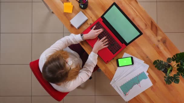 Primer plano de la sesión superior de la joven mujer de negocios caucásica utilizando el ordenador portátil con el teléfono con pantalla verde acostado en el escritorio en el interior de la oficina — Vídeo de stock
