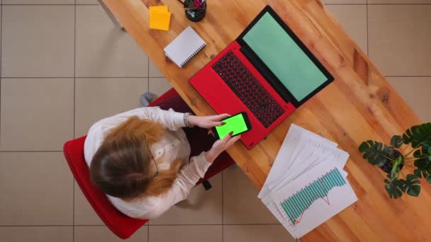 Close-up top shoot van jonge Kaukasische zakenvrouw het gebruik van de telefoon met groen scherm zittend in de voorkant van de laptop binnenshuis in het kantoor — Stockvideo