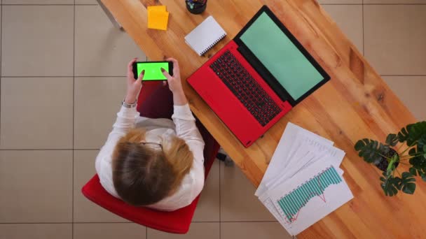 Κοντινά πλάνα της νεαρής καυκάσιας επιχειρηματίας παίζοντας βιντεοπαιχνίδια στο τηλέφωνο με πράσινη οθόνη κάθεται μπροστά από το φορητό υπολογιστή σε εσωτερικούς χώρους στο γραφείο — Αρχείο Βίντεο
