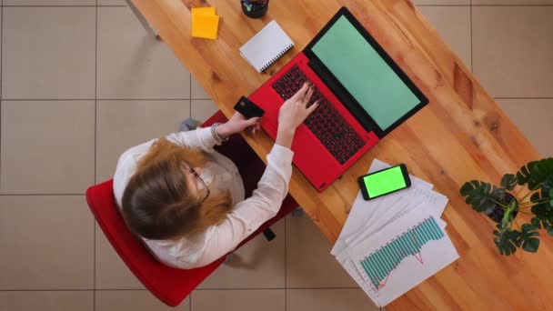 Nahaufnahme Top-Shooting der jungen kaukasischen Geschäftsfrau Online-Shopping mit der Kreditkarte auf dem Laptop drinnen mit Telefon mit grünem Bildschirm auf dem Schreibtisch im Büro liegend — Stockvideo