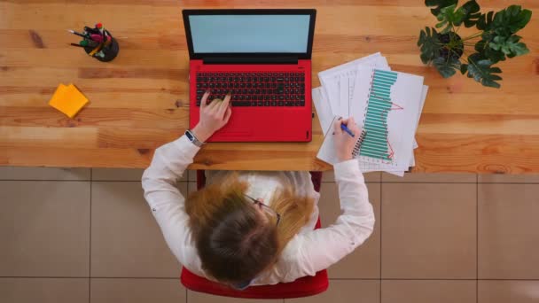 Närbild topp shoot av unga kaukasiska affärskvinna i glasögon med hjälp av den bärbara datorn och anteckningar inomhus på kontoret — Stockvideo