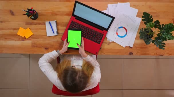 Close-up top shoot van jonge blanke werknemer met behulp van de Tablet met groen scherm zittend in de voorkant van de laptop binnenshuis in het kantoor — Stockvideo