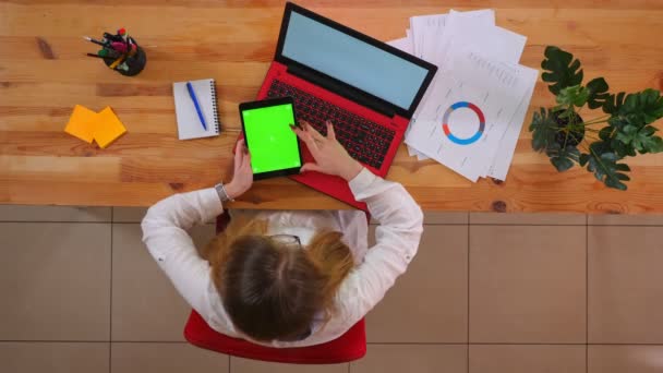 Close-up top shoot de jovem funcionário caucasiano usando o tablet com tela de croma verde sentado na frente do laptop dentro de casa no escritório — Vídeo de Stock