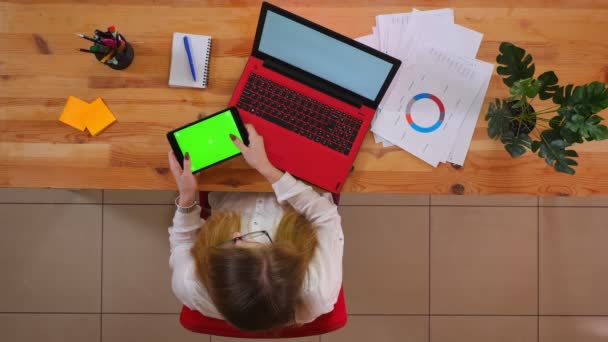 Close-up tiro superior de jovem funcionário caucasiano digitando no tablet com tela de croma verde sentado na frente do laptop dentro de casa no escritório — Vídeo de Stock