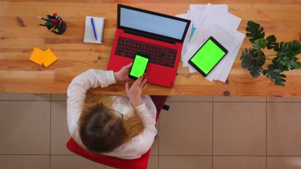 Close-up top shoot van jonge blanke werknemer met behulp van de telefoon met de Tablet met groen scherm liggend op het Bureau zittend voor de laptop binnenshuis in het kantoor — Stockvideo