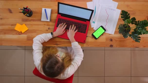 Primo piano ripresa superiore di giovane dipendente caucasico utilizzando il computer portatile con il telefono con schermo verde sdraiato sulla scrivania all'interno dell'ufficio — Video Stock