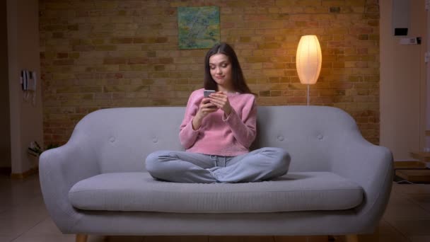 Съемки крупным планом молодой красивой брюнетки кавказской женщины с помощью телефона и радостно улыбающейся сидящей на диване в уютной квартире в помещении — стоковое видео