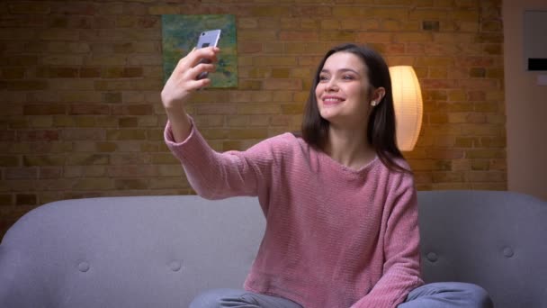 Primo piano ripresa di giovane bella bruna femmina caucasica scattare selfie al telefono e sorridente seduto sul divano in un accogliente appartamento al chiuso — Video Stock