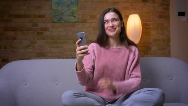 Närbild shoot av ung Söt brunett kaukasiska kvinna som har ett videosamtal på telefonen och leende sitter på soffan i en mysig lägenhet inomhus — Stockvideo