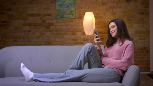 Close-up shoot van jonge mooie brunette Kaukasische vrouw met behulp van de telefoon kijken naar camera en glimlachend zittend op de Bank in een gezellig appartement binnenshuis — Stockvideo