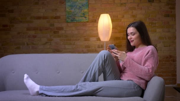 휴대 전화를 사용하여 젊은 예쁜 갈색 머리 백인 여성의 클로즈업 촬영 과 미소 앉아 누워에 소파에 아늑한 아파트 실내 — 비디오