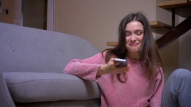 Telefonu kullanarak ve kapalı rahat bir dairede yerde oturan gülümseyen genç güzel esmer kafkas kadın Closeup ateş