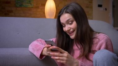 Telefonu kullanarak ve kapalı rahat bir dairede yerde oturan gülümseyen genç mutlu esmer kafkas kadın Closeup ateş