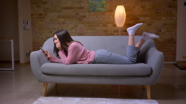Closeup hajtás fiatal csinos barna kaukázusi nő a telefon segítségével, és mosolyogva feküdt a kanapén egy hangulatos lakásban beltéri — Stock videók