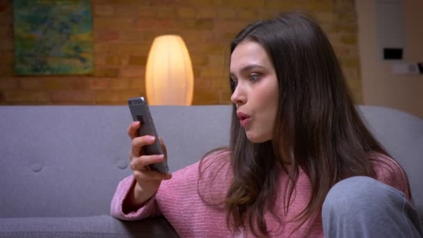 Close-up zijaanzicht schieten van jonge gelukkige brunette Kaukasische vrouw met behulp van de telefoon en glimlachend zittend op de vloer in een gezellig appartement binnenshuis — Stockvideo