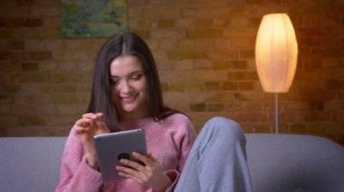 Tablet kullanarak genç güzel esmer kafkas kadın Closeup ateş ve kapalı rahat bir dairede kanepede oturan gülümseyerek
