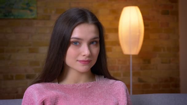 Съемки крупным планом юной брюнетки кавказской девушки, смотрящей в камеру и счастливо улыбающейся сидящей на диване в уютной квартире в помещении — стоковое видео
