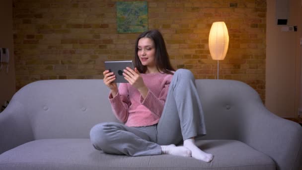 Съемки крупным планом молодой симпатичной брюнетки кавказской женщины, переписывающейся на планшете, сидящей на диване в уютной квартире в помещении — стоковое видео