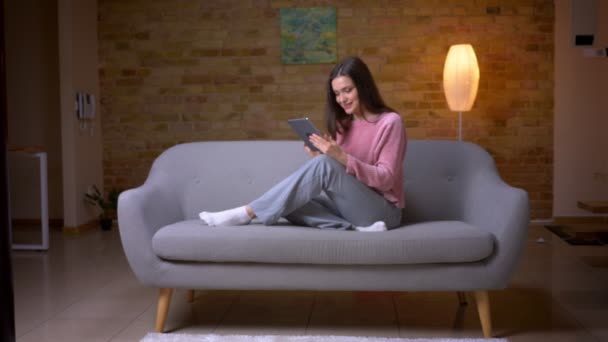 Съемки крупным планом молодой красивой брюнетки кавказской женщины, переписывающейся на планшете и счастливо улыбающейся, сидящей на диване в уютной квартире в помещении — стоковое видео
