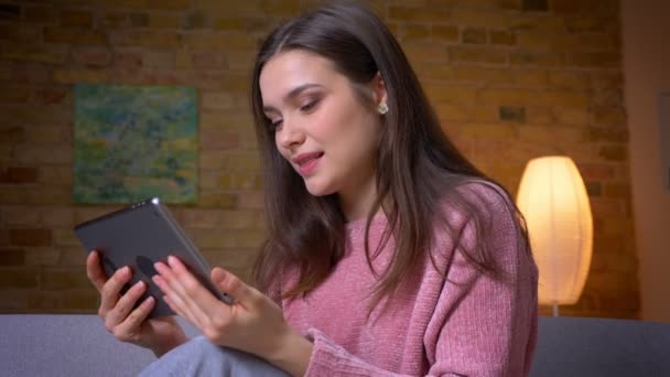Nahaufnahme einer jungen hübschen brünetten kaukasischen Frau, die auf dem Tablet surft und glücklich in einer gemütlichen Wohnung drinnen lächelt — Stockvideo