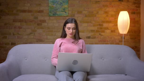 Крупним планом знімок молодої красивої брюнетки біла жінка використовує ноутбук і посміхається сидячи на дивані в затишній квартирі в приміщенні — стокове відео