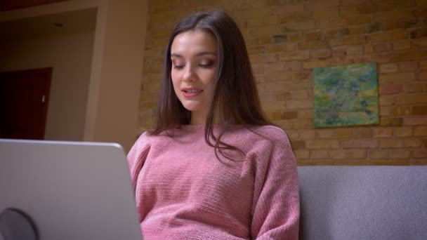 Primer plano de la joven guapa morena caucásica mujer de negocios trabajando en el portátil y sonriendo sentado en el sofá en un acogedor apartamento en el interior — Vídeo de stock