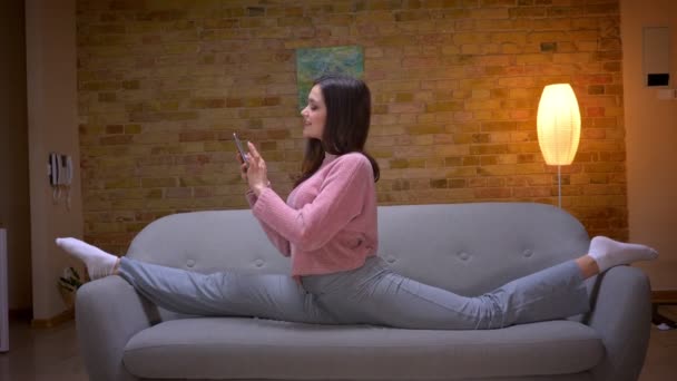 Gros plan shoot de jeune jolie brune caucasienne en utilisant le téléphone etassis dans la ficelle sur le canapé dans un appartement confortable à l'intérieur — Video