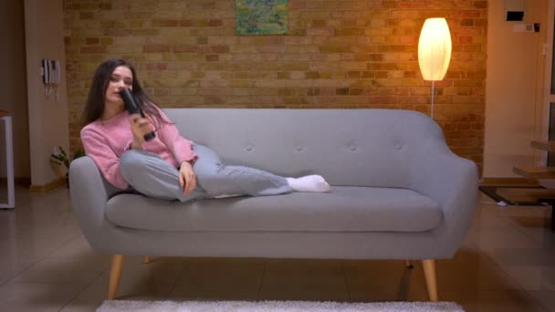 Nahaufnahme einer jungen hübschen brünetten kaukasischen Frau, die eine Komödie im Fernsehen anschaut und lacht, wenn sie auf der Couch in einer gemütlichen Wohnung drinnen sitzt — Stockvideo