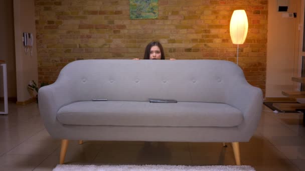 Gros plan de jeune jolie brune caucasienne regardant un film d'horreur à la télévision et se cachant derrière le canapé dans un appartement confortable à l'intérieur — Video