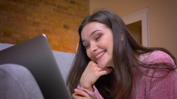 Primo piano ripresa di giovane bella bruna adolescente caucasica utilizzando il computer portatile e ridendo felicemente sul divano in un accogliente appartamento al chiuso — Video Stock