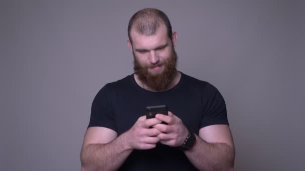 Съемка крупным планом взрослого красивого мускулистого белого мужчину с бородой с помощью телефона перед камерой на фоне изолированного на сером фоне — стоковое видео