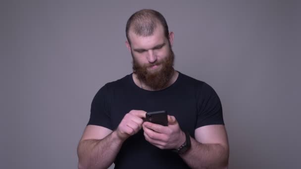 Gros plan shoot de adulte beau musclé caucasien homme avec barbe messagerie sur le téléphone en face de la caméra avec fond isolé sur gris — Video