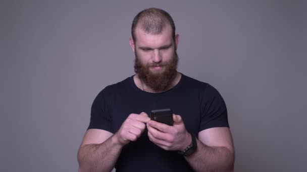 Съемка крупным планом взрослого красивого мускулистого белого мужчину с бородой, печатающего по телефону перед камерой на фоне изолированного на сером фоне — стоковое видео