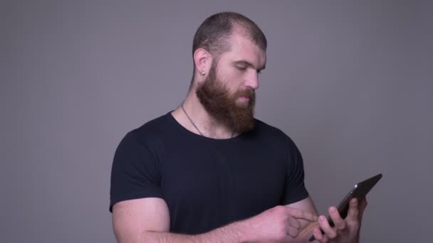 Primer plano del hombre caucásico musculoso guapo adulto con barba usando la tableta delante de la cámara con fondo aislado en gris — Vídeo de stock