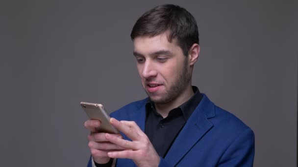 Gros plan pousse de jeune homme caucasien beau en utilisant le téléphone en regardant la caméra et souriant avec fond isolé sur gris — Video