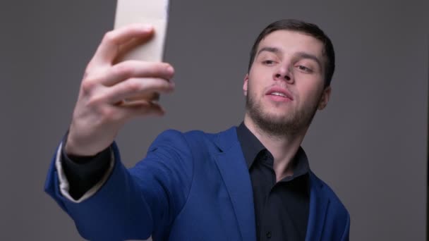 Primer plano disparar de joven guapo caucásico hombre tomando selfies en el teléfono con fondo aislado en gris — Vídeo de stock