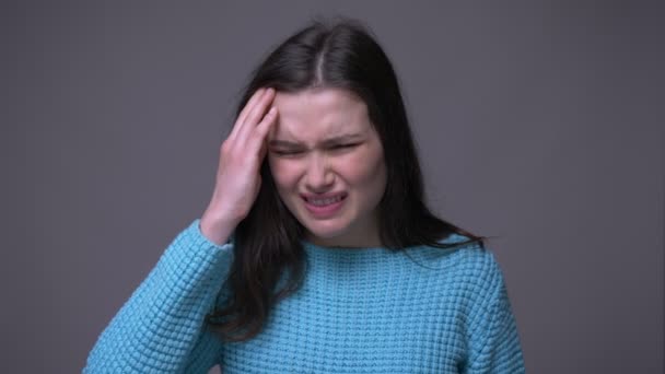 Nahaufnahme einer jungen hübschen brünetten Frau mit Kopfschmerzen vor der Kamera mit isoliertem grauem Hintergrund — Stockvideo