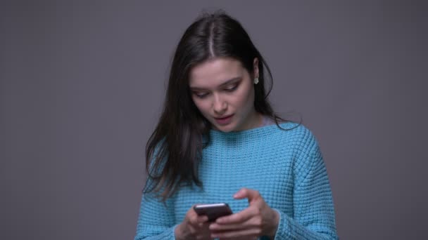 Gros plan tige de jeune jolie brune femelle en utilisant le téléphone et souriant avec fond isolé sur gris — Video