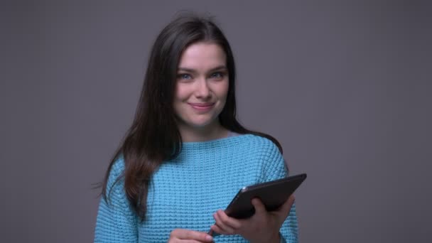 Close-up tiro de jovem morena bonita do sexo feminino usando o tablet e mostrando tela verde olhando para a câmera com fundo isolado em cinza — Vídeo de Stock