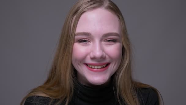 Крупный план портрета юной привлекательной брюнетки, улыбающейся весело глядя в камеру с изолированным на сером фоне — стоковое видео
