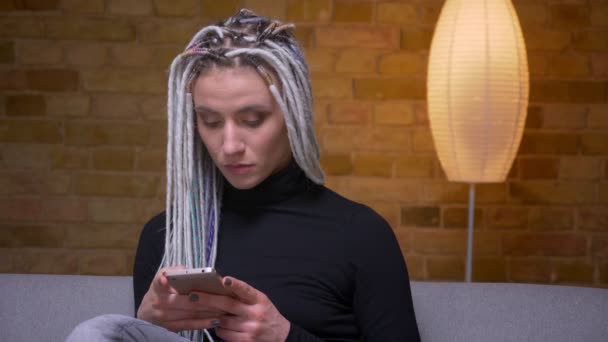 Zbliżenie strzelać młodych atrakcyjne kaukaski Hipster kobiet z blond dredy za pomocą telefonu uśmiechnięty szczęśliwie siedząc na kanapie w pomieszczeniu — Wideo stockowe